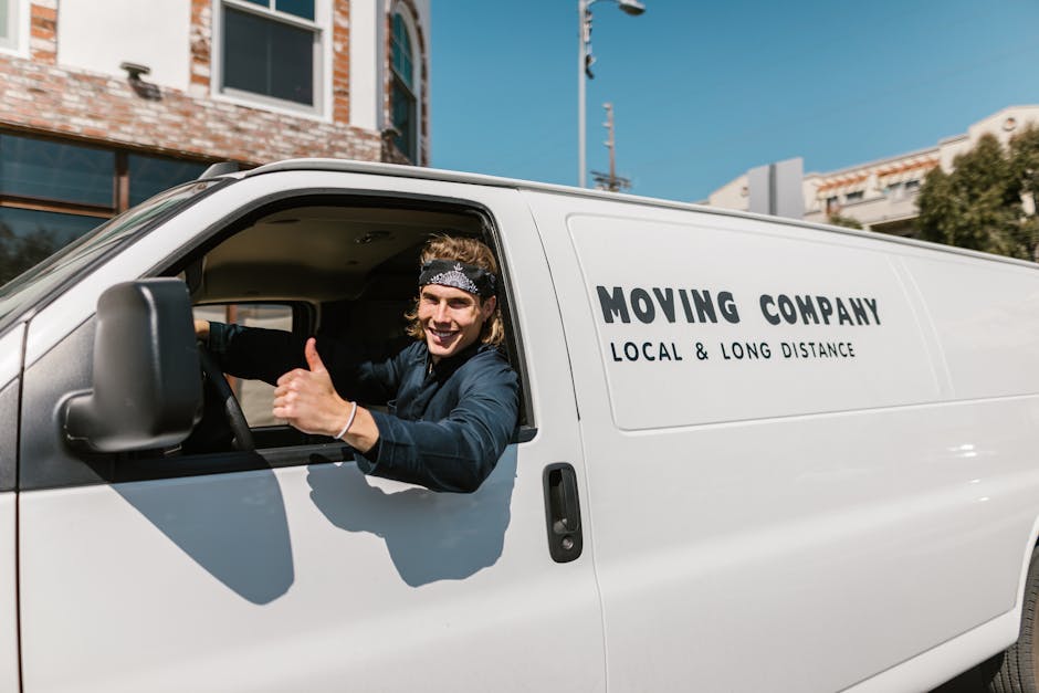 A young male mover with shoulder-length hair and a black bandana, smiling and giving a thumbs-up gesture while seated in the driver's seat of a white moving van with the words 'MOVING COMPANY LOCAL & 